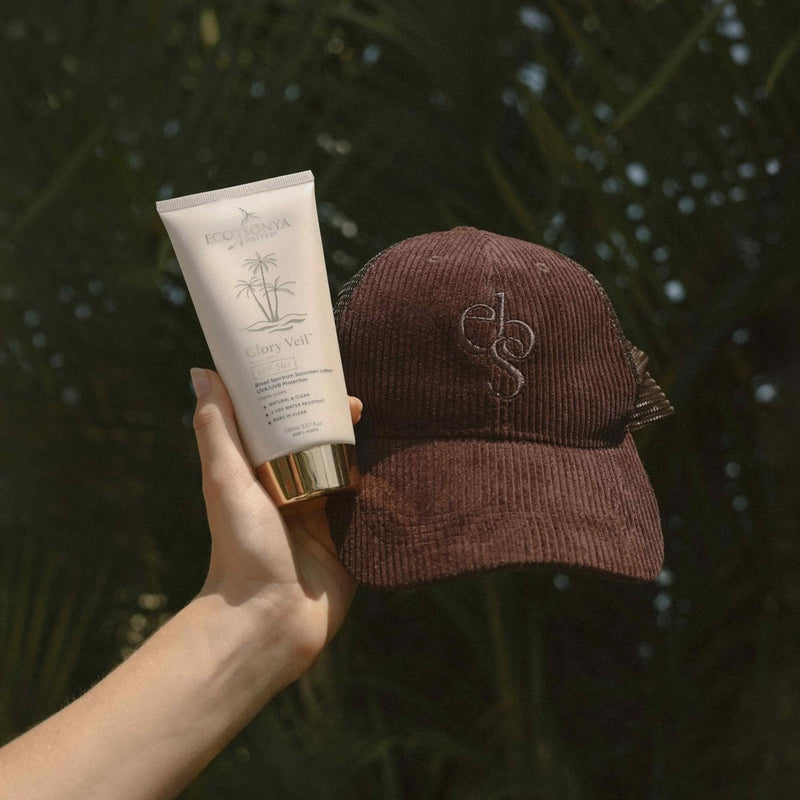 Hand holding Eco By Sonya Glory Veil sunscreen alongside the Good Thoughts Cap on a white background.