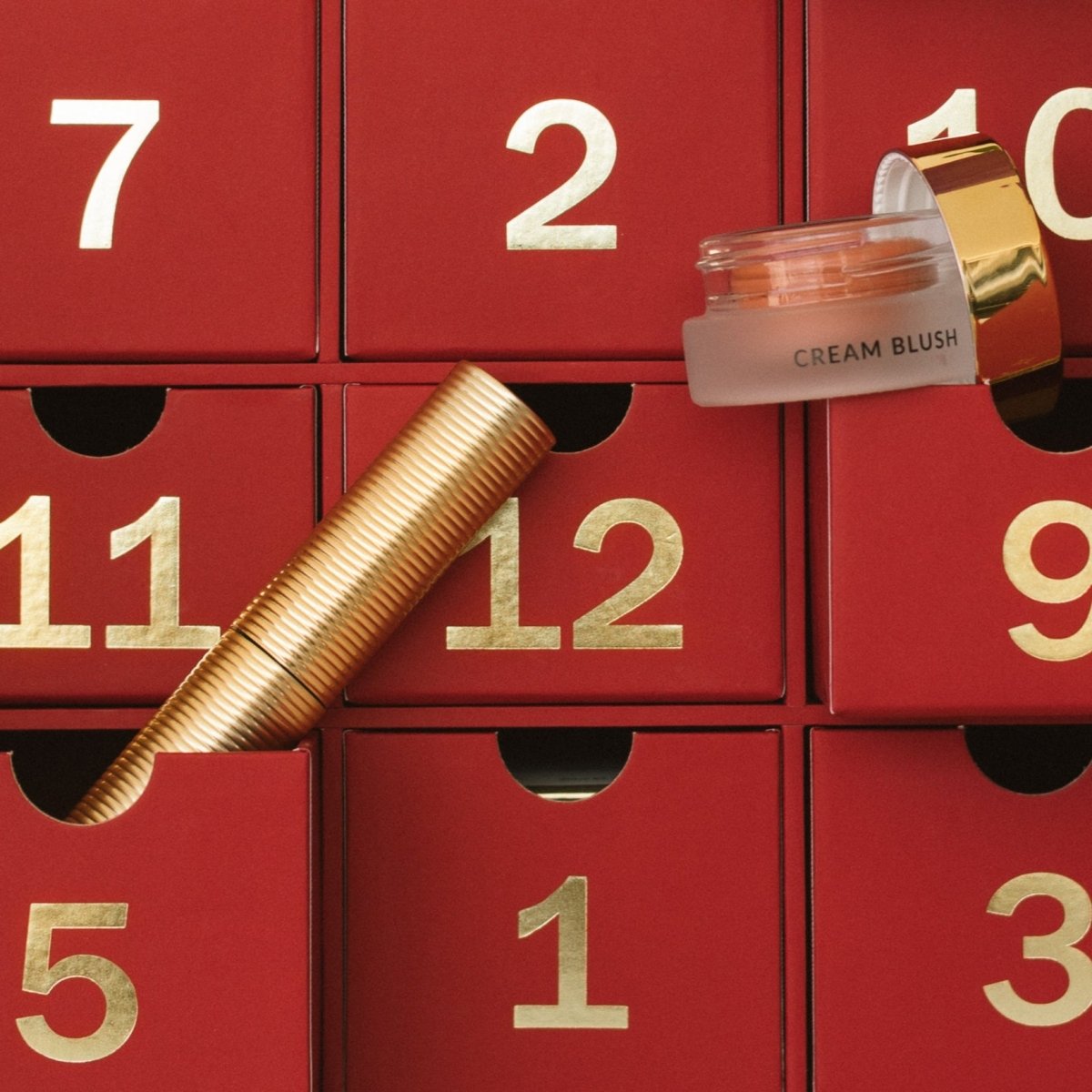 Red advent calendar with gold numbered drawers, featuring a cream blush and gold tube mascara.