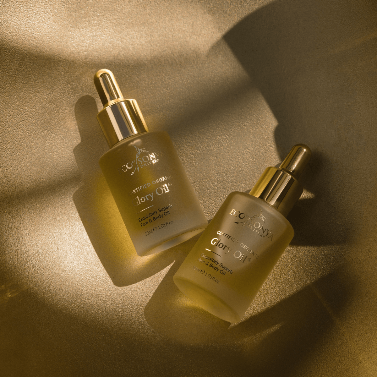 Two Eco By Sonya Glory Oil bottles with golden droppers on golden textured surface, softly lit by sunlight for a warm natural glow.
