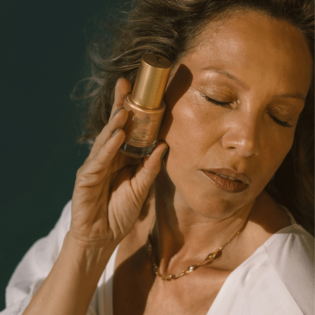 Mature woman with radiant golden skin holding Eco By Sonya Golden Illuminiser bottle near her face, showing luminous youthful glow and warm sunlight.
