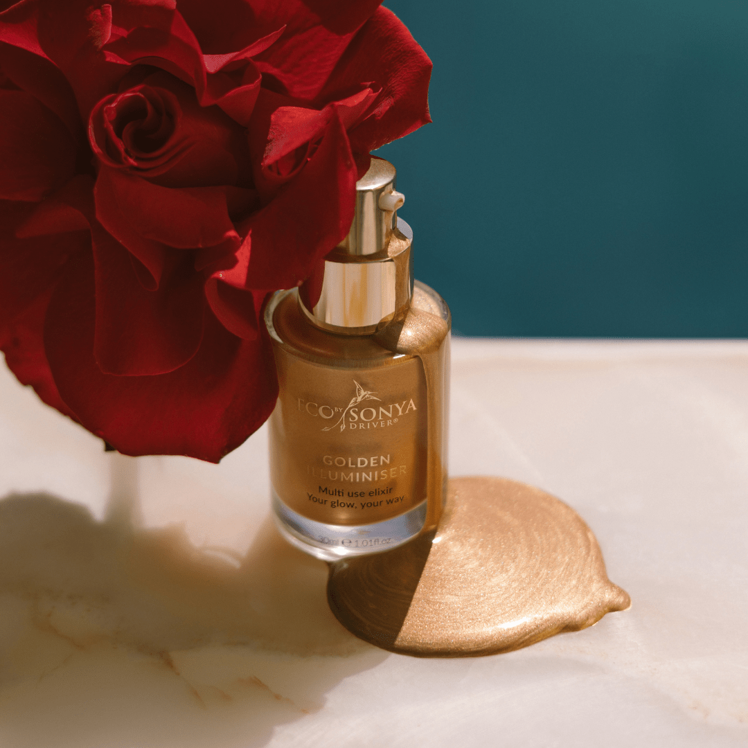Eco By Sonya Golden Illuminiser bottle with golden product dripping beside red rose on marble surface, glowing liquid texture.