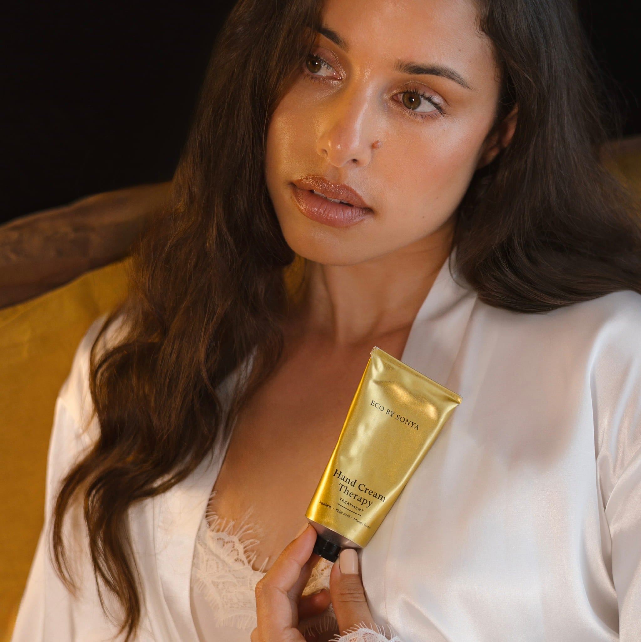 Woman in a white satin robe holding a gold tube of Eco By Sonya Hand Cream Therapy Treatment against a dark background.