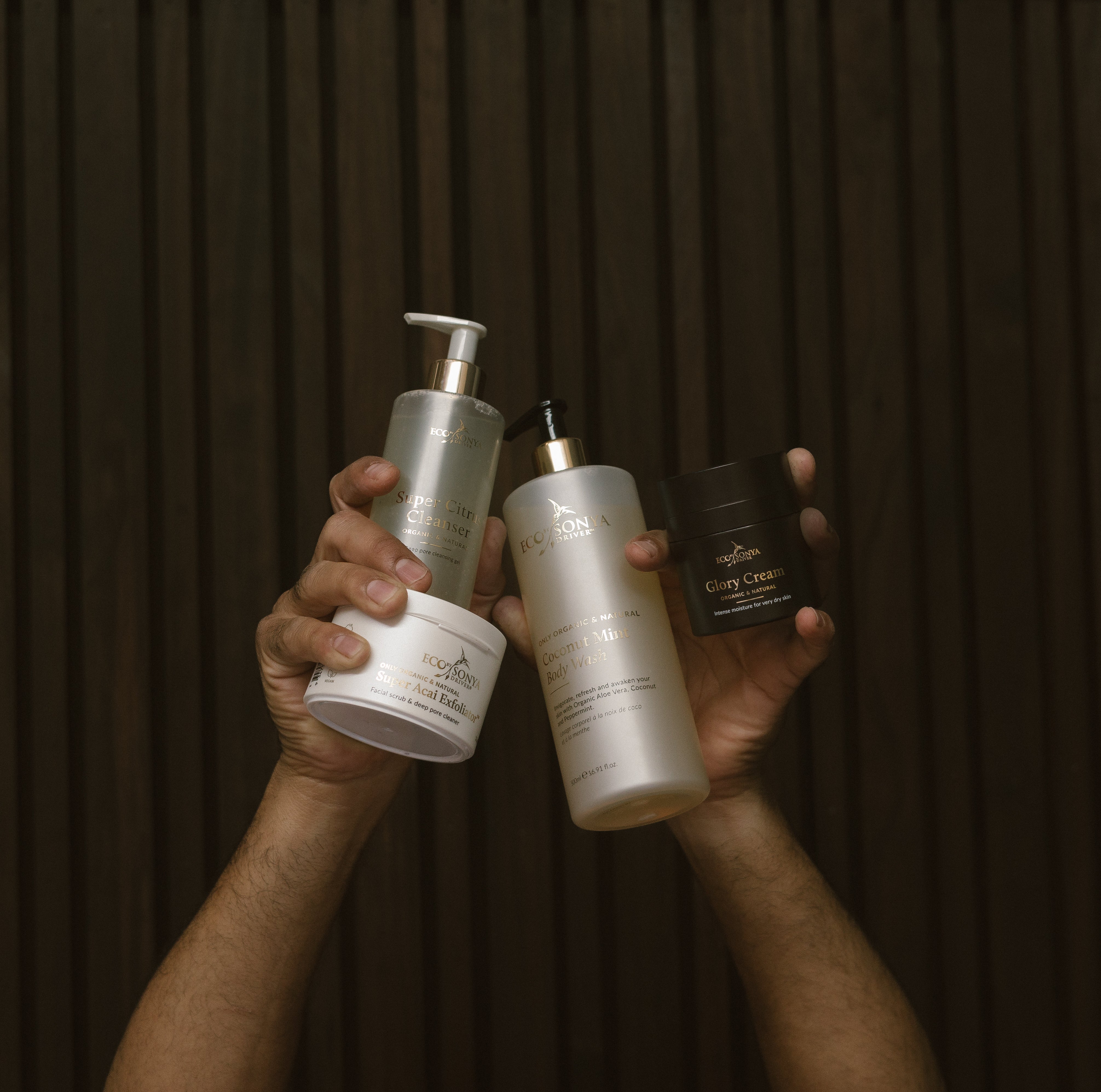 Hands holding various Eco By Sonya skincare products from Mature Blokes Pack against a dark background