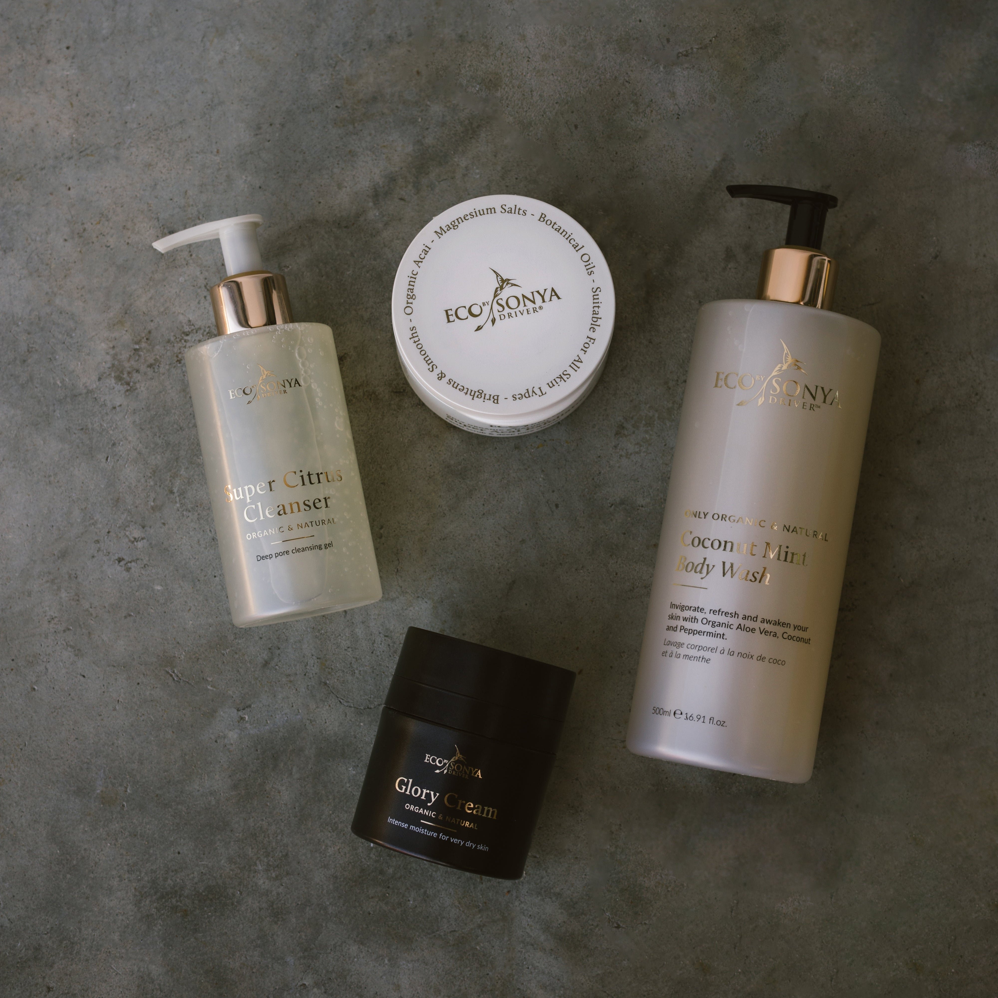 Set of Eco By Sonya skincare products including a Super Citrus Cleanser, Acai Exfoliator, Glory Cream and Coconut Mint Body Wash in a gray surface.