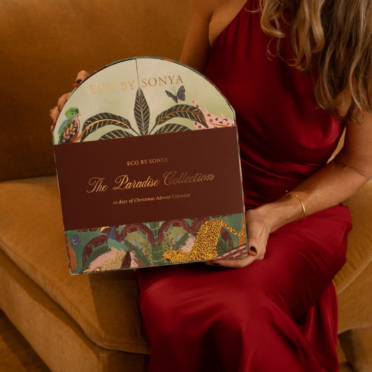 Woman in a red satin dress holding the Eco By Sonya The Paradise Collection Christmas Advent Calendar while seated on a couch.