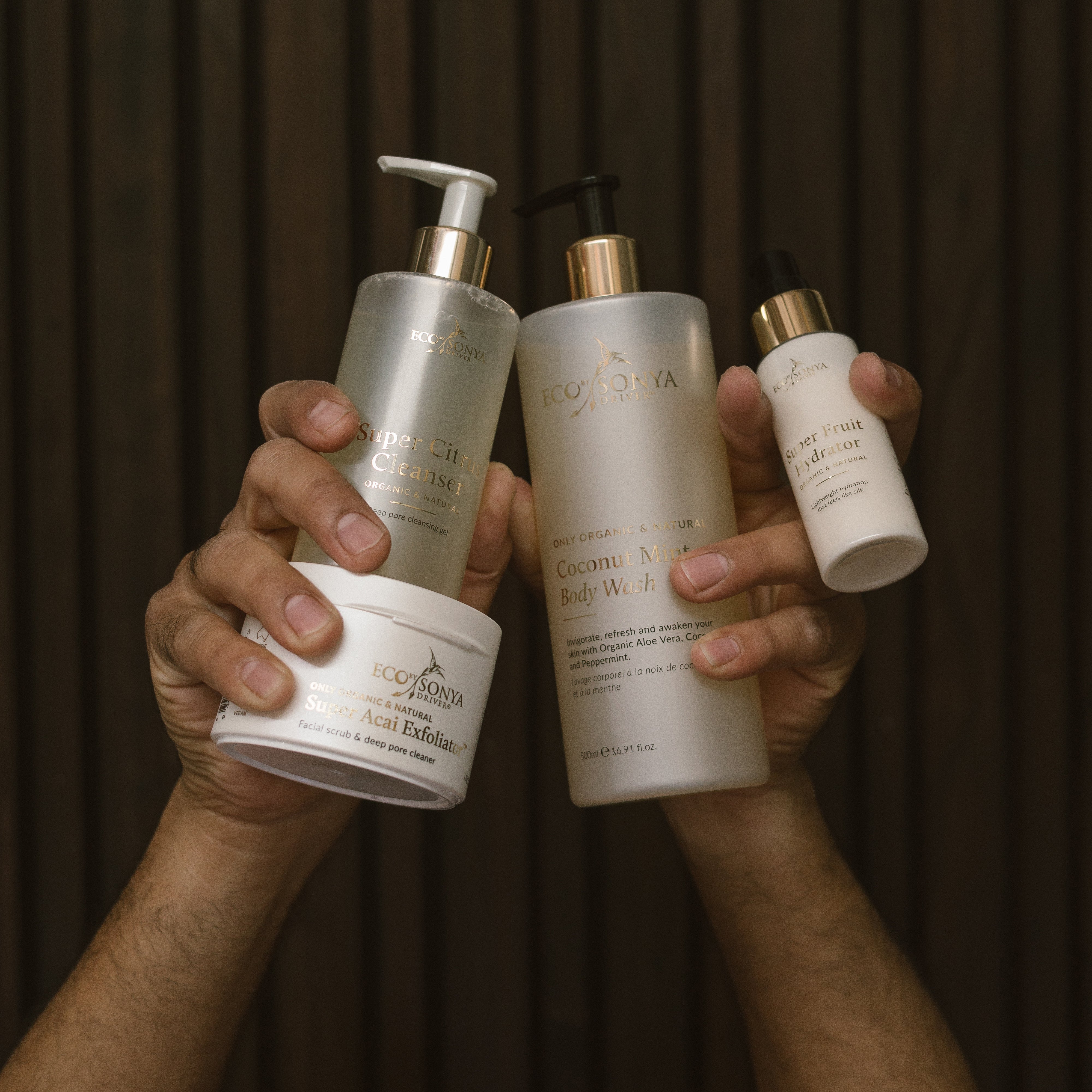 Hands holding various Eco By Sonya skincare products from Young Blokes Pack against a dark background