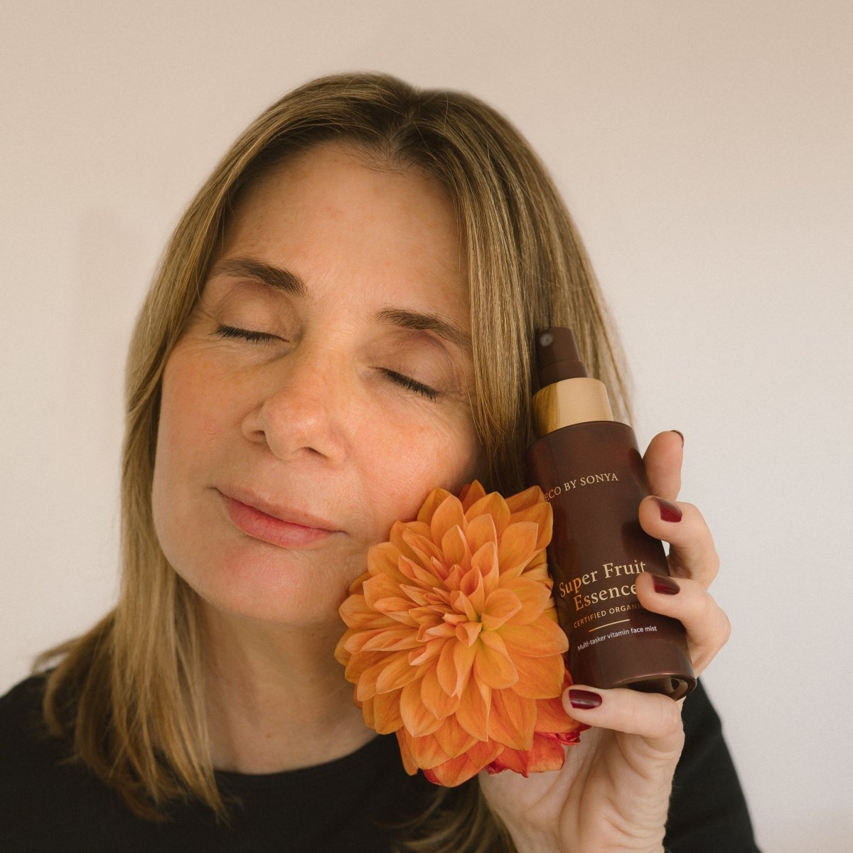 Woman holding an orange flower and a bottle of Eco By Sonya Super Fruit Essence against a plain background
