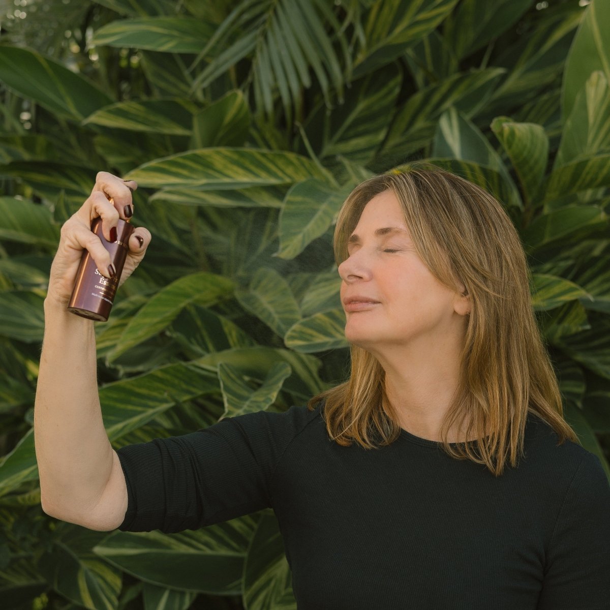 Woman spraying Eco By Sonya Super Fruit Essence onto her face, against a green leafy background